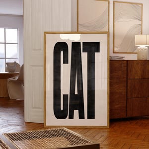 May include: A framed art print with the word "CAT" in large, bold, black letters against a cream background. The print is in a light wood frame and is displayed on a wooden floor. Other framed art pieces are visible in the background.