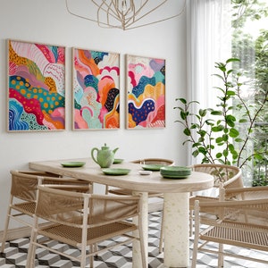 May include: Three abstract paintings with colorful, swirling patterns in shades of pink, blue, yellow, green, and orange hang on a white wall above a wooden dining table with four chairs.