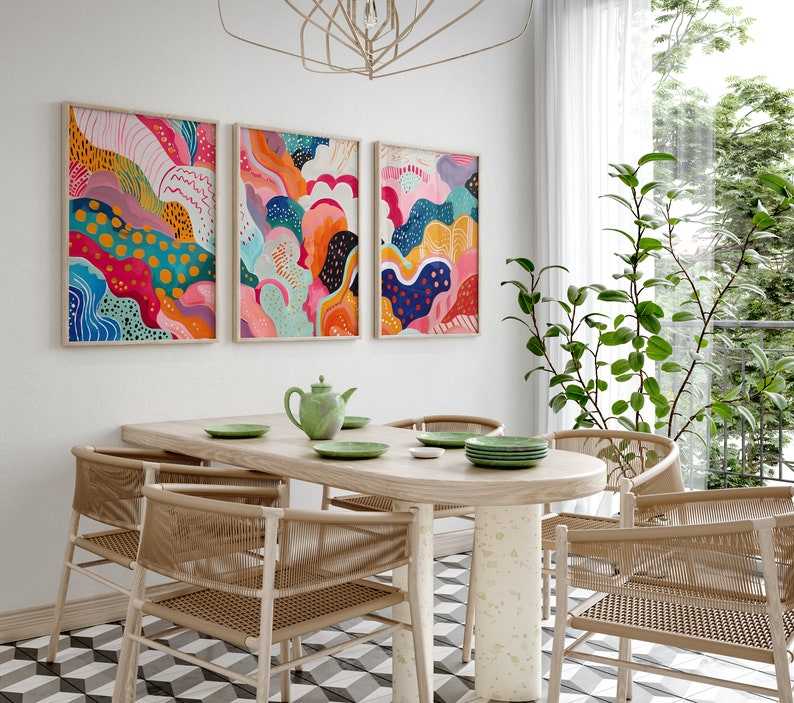 May include: Three abstract paintings with colorful, swirling patterns in shades of pink, blue, yellow, green, and orange hang on a white wall above a wooden dining table with four chairs.