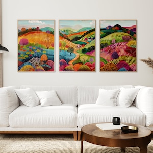 Może przedstawiać: Trzy oprawione obrazy przedstawiające krajobrazy z kolorowymi wzgórzami, drzewami i krętym szlakiem. Obrazy znajdują się w nowoczesnym salonie z białą sofą, stolikiem kawowym i wazonem z suszonymi kwiatami.