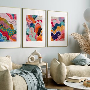 May include: Three framed abstract paintings with colorful, swirling patterns in shades of pink, blue, green, yellow, and orange. The paintings are hung on a light blue wall in a living room setting.