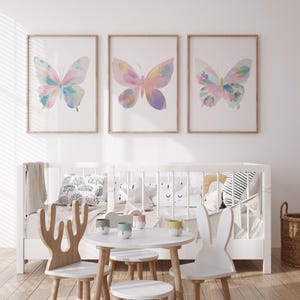 Op de afbeelding: Drie ingelijste aquarelprints van vlinders in pastelkleuren hangen boven een wit babybedje. De vlinders hebben tinten roze, blauw en geel. Een kleine witte tafel en stoelen staan voor het bedje, wat een grillige kinderkameromgeving creëert.