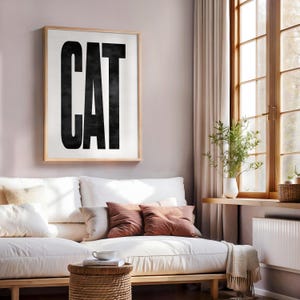 May include: A framed art print with the word "CAT" in bold, black letters against a white background. The artwork is displayed above a white sofa with brown throw pillows. The room has a window with natural light.