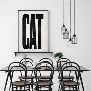 May include: A dining room scene featuring a dark wood table with six black chairs with woven seats. Above the table hang three black cage pendant lights. A framed print on the wall reads "CAT" in bold black letters.
