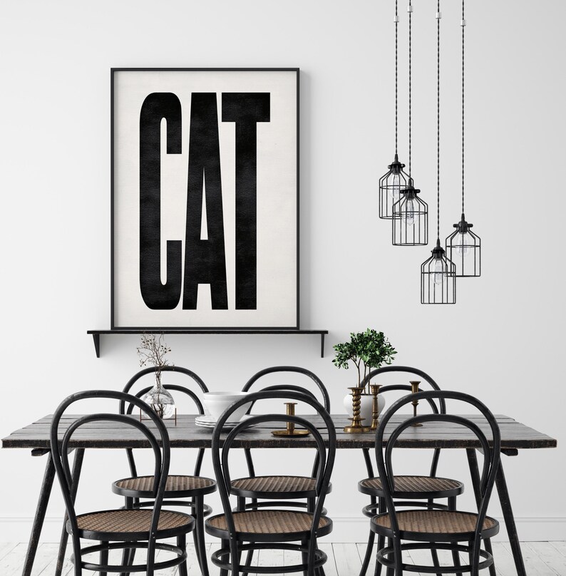 May include: A dining room scene featuring a dark wood table with six black chairs with woven seats. Above the table hang three black cage pendant lights. A framed print on the wall reads "CAT" in bold black letters.