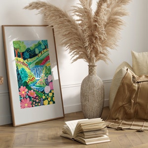 May include: A framed print of a colorful painting depicting a lush green landscape with a waterfall, flowers, and a winding stream. The print is displayed on a wooden floor with a book and a vase of pampas grass.