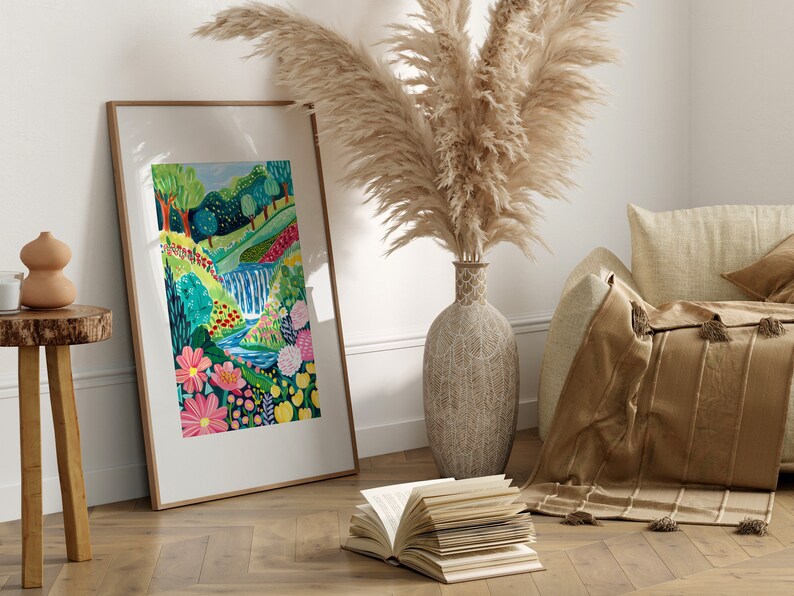 May include: A framed print of a colorful painting depicting a lush green landscape with a waterfall, flowers, and a winding stream. The print is displayed on a wooden floor with a book and a vase of pampas grass.