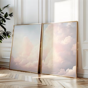 May include: Two framed prints depicting a pastel pink and blue sky with fluffy white clouds. The prints are in gold frames and are leaning against a white wall.