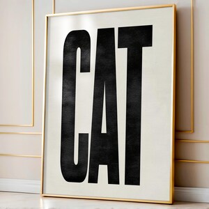 May include: A framed art print featuring the word "CAT" in bold, black, uppercase letters against a cream-colored background. The frame is a thin, gold color. The artwork is displayed in a room with gold trim.