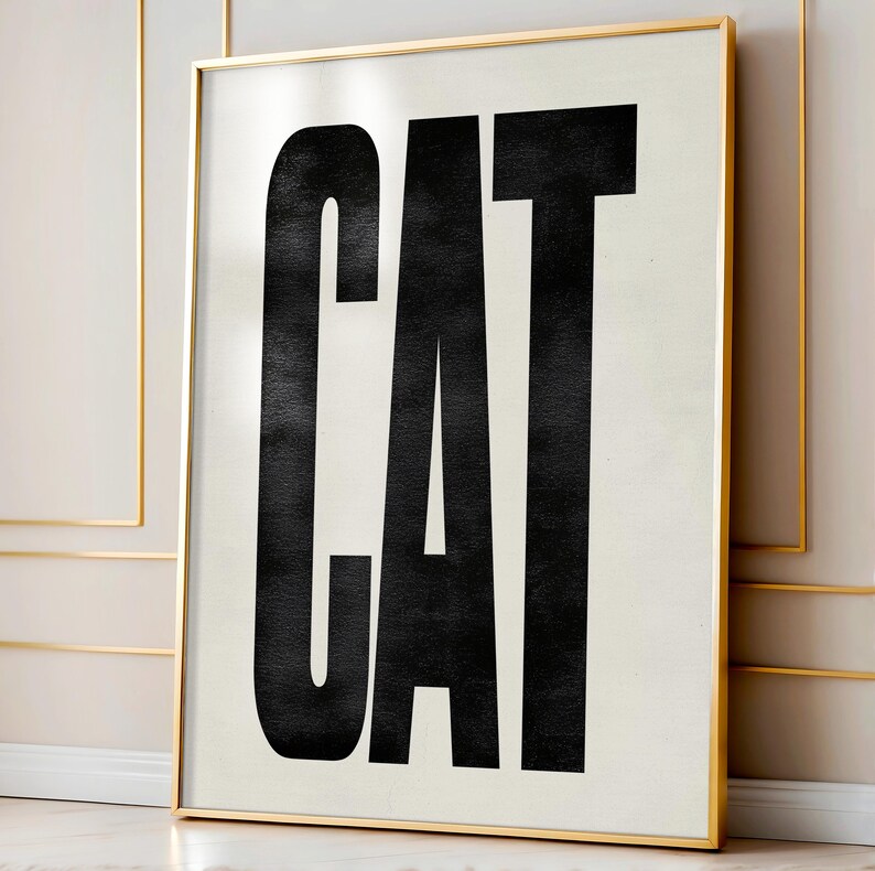 May include: A framed art print featuring the word "CAT" in bold, black, uppercase letters against a cream-colored background. The frame is a thin, gold color. The artwork is displayed in a room with gold trim.