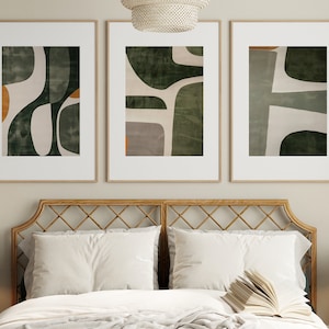 Op de afbeelding: Drie ingelijste abstracte kunstprints met een groen, wit en oranje kleurenschema. De prints hangen aan een muur boven een houten hoofdeinde.