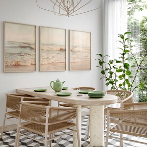 May include: A dining room with a wooden table and six woven chairs. The table has a green teapot and four plates on it. Three framed pictures of ocean waves hang on the wall. A potted plant is in the corner of the room.