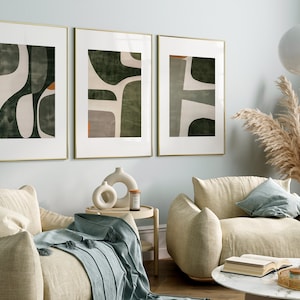 Op de afbeelding: Drie ingelijste abstracte kunstprints met een groen, wit en oranje kleurenschema hangen aan een lichtblauwe muur boven twee beige fauteuils. De fauteuils staan in een woonkamer met een bijzettafel, een salontafel en een vaas met gedroogd pampasgras.