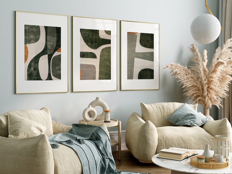 Op de afbeelding: Drie ingelijste abstracte kunstprints met een groen, wit en oranje kleurenschema hangen aan een lichtblauwe muur boven twee beige fauteuils. De fauteuils staan in een woonkamer met een bijzettafel, een salontafel en een vaas met gedroogd pampasgras.