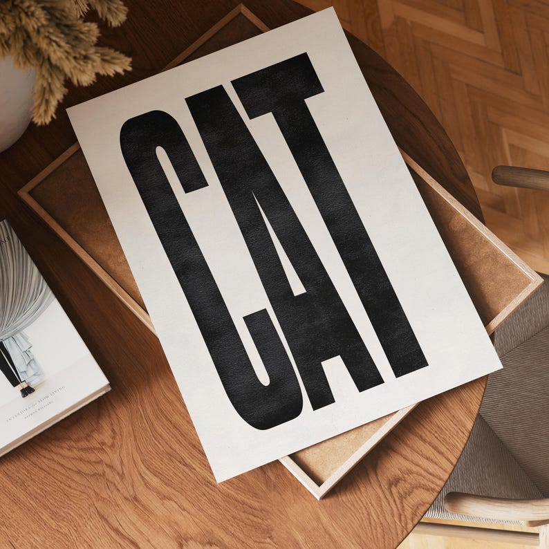 May include: A print with the word "CAT" in large, bold, black letters. The print is on a cream-colored background and is displayed on a wooden table. The print is inside a wooden frame.
