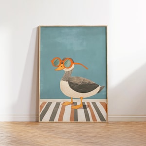 May include: Framed art print featuring a whimsical illustration of a duck wearing orange-rimmed glasses. The duck is depicted with gray and white feathers, standing on a striped rug against a teal background. The artwork is framed in a light wood.