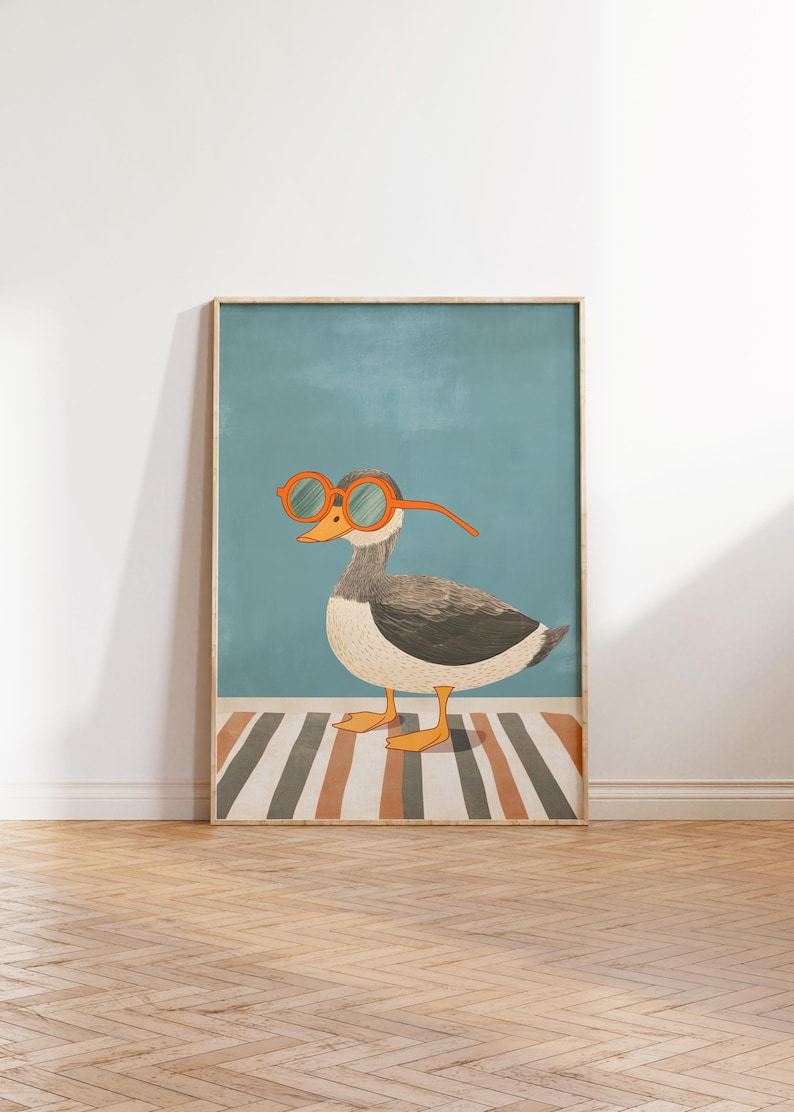 May include: Framed art print featuring a whimsical illustration of a duck wearing orange-rimmed glasses. The duck is depicted with gray and white feathers, standing on a striped rug against a teal background. The artwork is framed in a light wood.