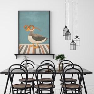 May include: A framed art print featuring a whimsical illustration of a goose wearing orange glasses. The goose stands on a striped surface against a teal background. The print is displayed above a black dining table with black chairs.