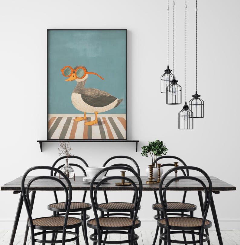 May include: A framed art print featuring a whimsical illustration of a goose wearing orange glasses. The goose stands on a striped surface against a teal background. The print is displayed above a black dining table with black chairs.