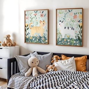 Puede incluir: Dos impresiones enmarcadas de arte de gatos con marcos de madera cuelgan sobre una cama con ropa de cama y almohadas a rayas. Las impresiones presentan gatos en un jardín floral. Una cómoda blanca con animales de peluche se encuentra a la izquierda.