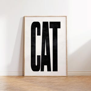 May include: Framed art print featuring the word "CAT" in bold, black capital letters against a white background. The print is in a light wood frame, set against a white wall and a wooden floor.