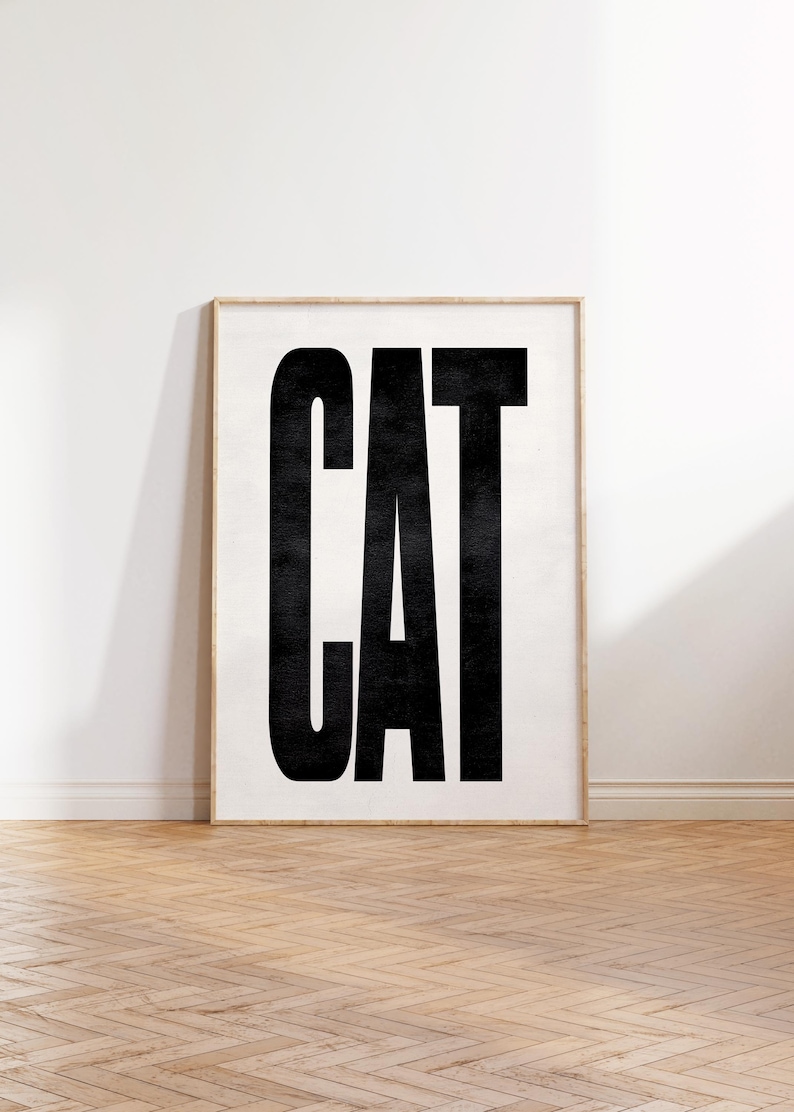 May include: Framed art print featuring the word "CAT" in bold, black capital letters against a white background. The print is in a light wood frame, set against a white wall and a wooden floor.