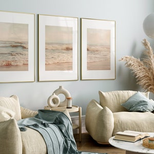 May include: Three framed prints of a beach scene with waves crashing on the shore. The prints are in gold frames and are hung on a light blue wall. The prints are in a living room setting with two beige armchairs and a coffee table.