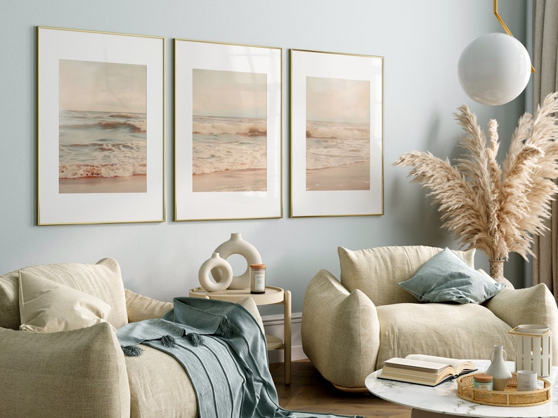 May include: Three framed prints of a beach scene with waves crashing on the shore. The prints are in gold frames and are hung on a light blue wall. The prints are in a living room setting with two beige armchairs and a coffee table.
