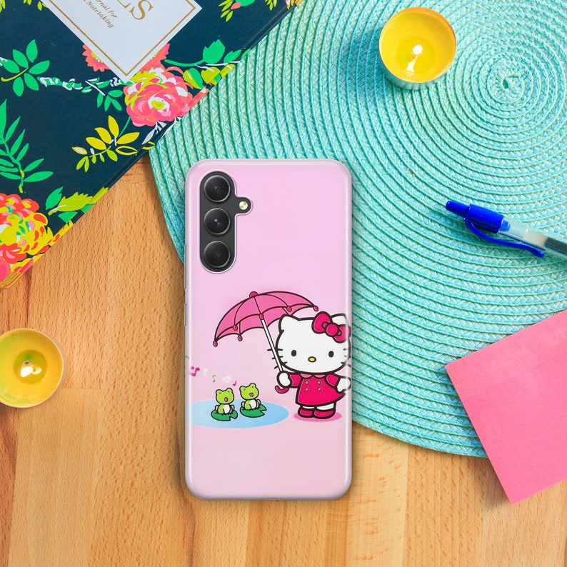 Puede incluir: Funda de tel&eacute;fono rosa con un dise&ntilde;o de Hello Kitty. Hello Kitty sostiene un paraguas rosa y hay dos ranas verdes en el fondo.