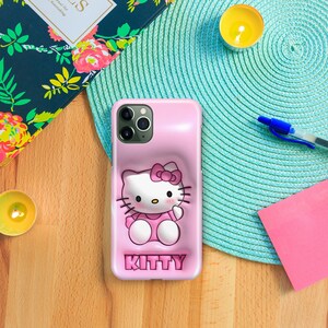 以下が含まれることがあります： 3Dのハローキティのデザインが施されたピンクの携帯電話ケース。ケースには、立体的なハローキティのフィギュアと、下部に「KITTY」という文字が印刷されています。