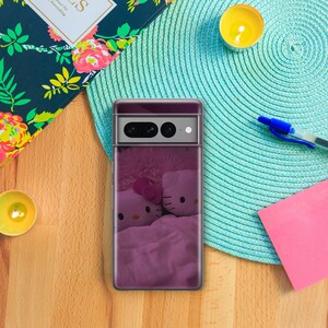May include: A clear phone case with a purple and pink design showing two Hello Kitty plush toys under a blanket. The phone case is on a wooden surface with a blue placemat, a notebook, a pen, and candles.