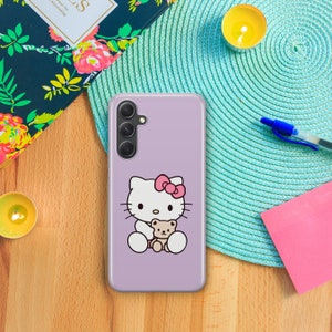 以下が含まれることがあります： ラベンダー色の携帯電話ケースに、白いハローキティのキャラクターが茶色のぬいぐるみを抱えています。携帯電話ケースには、3つのレンズが付いたカメラの切り抜きがあります。