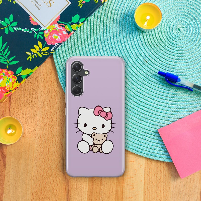 以下が含まれることがあります： ラベンダー色の携帯電話ケースに、白いハローキティのキャラクターが茶色のぬいぐるみを抱えています。携帯電話ケースには、3つのレンズが付いたカメラの切り抜きがあります。