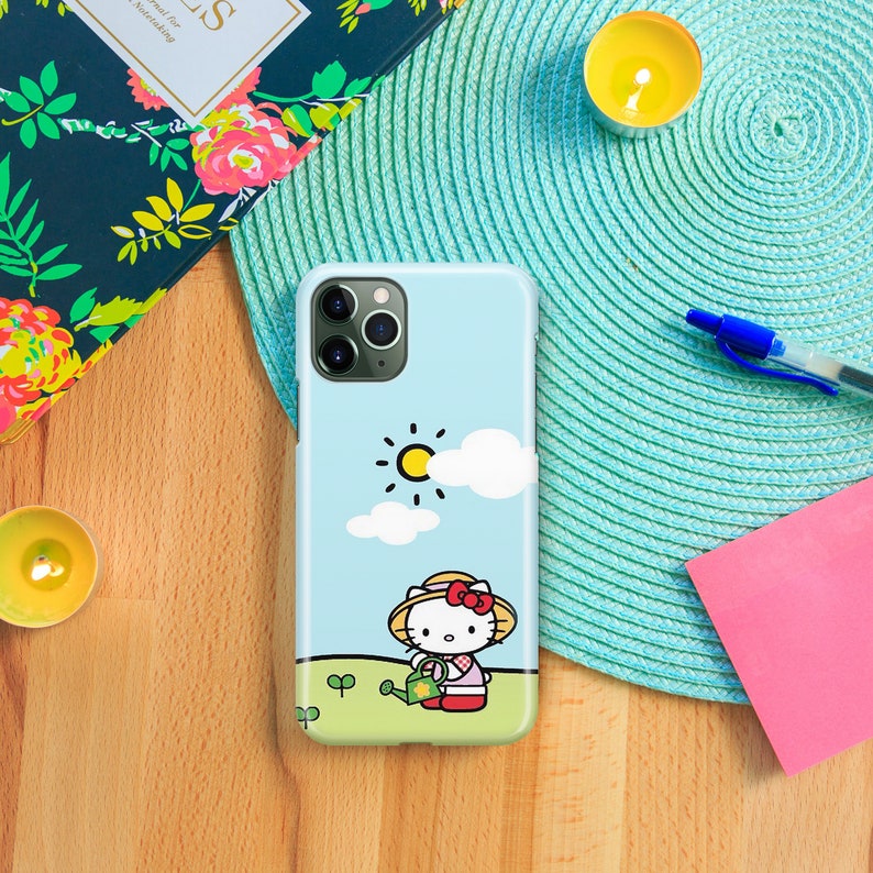 Pu&ograve; includere: Una custodia per telefono blu con un'immagine di Hello Kitty dei cartoni animati che innaffia le piante in un campo soleggiato. La custodia ha un ritaglio per la fotocamera del telefono.