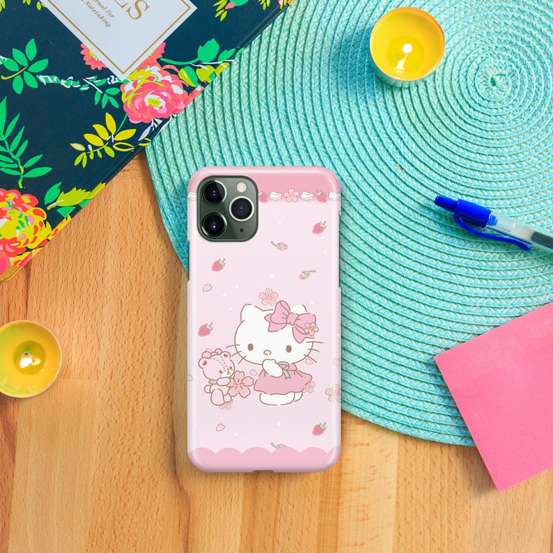 May include: A pink phone case with a white Hello Kitty design. Hello Kitty is holding a pink teddy bear. The case has a white border with pink flowers and strawberries.
