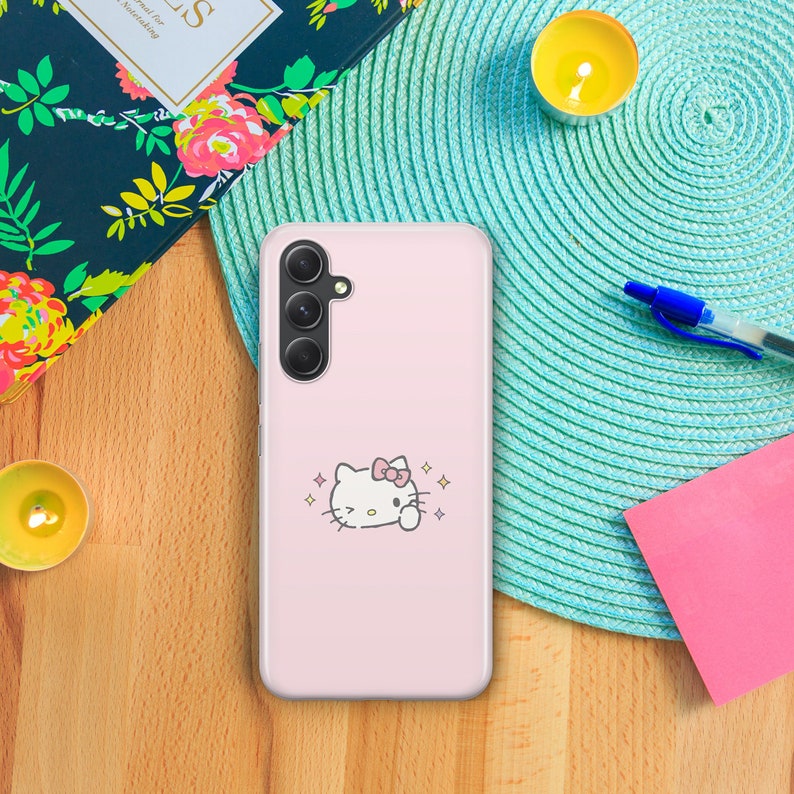 May include: A pink phone case with a white Hello Kitty design. The case has a camera cutout and a pink background.