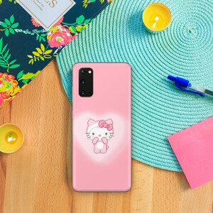 以下が含まれることがあります： ピンクの携帯電話ケースにハローキティのデザインが施されています。ケースはハローキティのキャラクターの周りに白いアウトラインがあり、ピンクの背景になっています。
