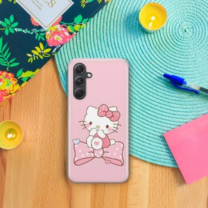以下が含まれることがあります： ピンクの携帯電話ケースに、ピンクのリボンをつけた白いハローキティのキャラクターが、白のドット柄のピンクのリボンに座っています。携帯電話ケースには、3つのレンズが付いたカメラの切り抜きがあります。