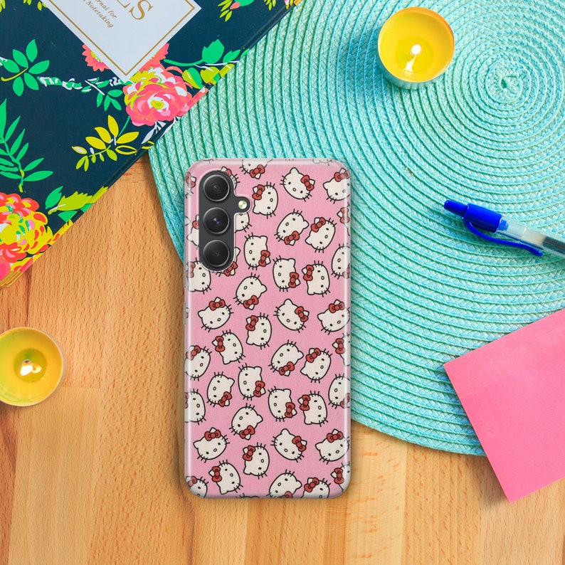 Op de afbeelding: Een roze telefoonhoesje met een herhalend patroon van Hello Kitty-gezichten. Het hoesje heeft een camera-uitsparing en een zwarte camera lens.