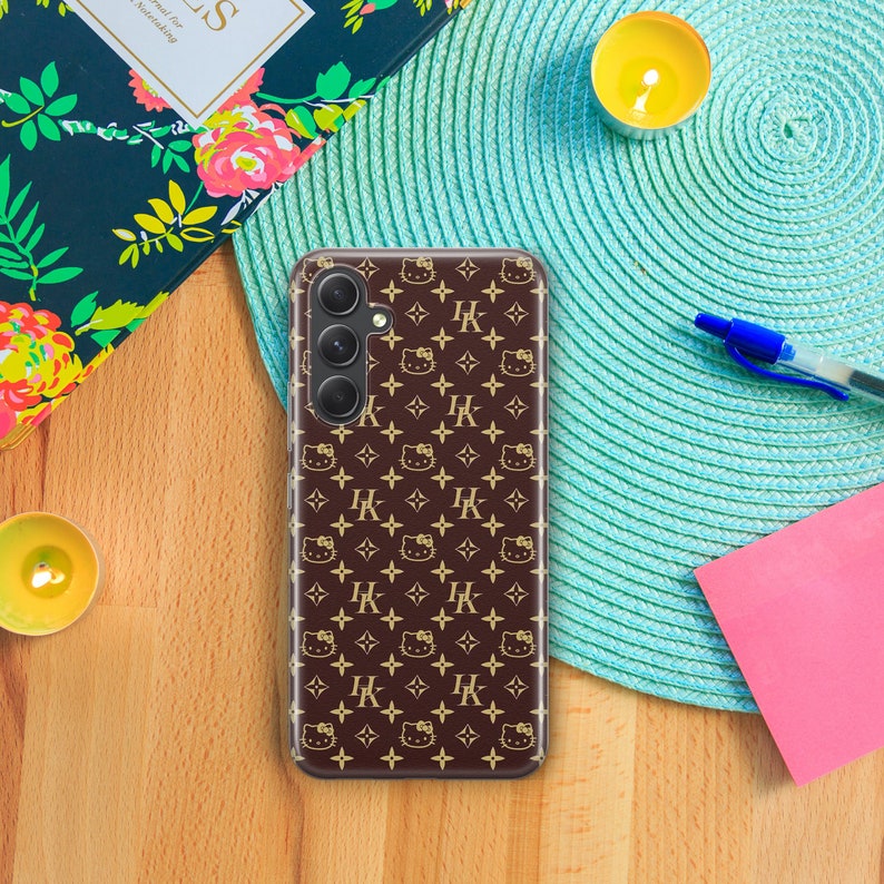 Op de afbeelding: Een bruine telefoonhoes met een gouden patroon van in elkaar grijpende 'HK'-letters en Hello Kitty-gezichten.