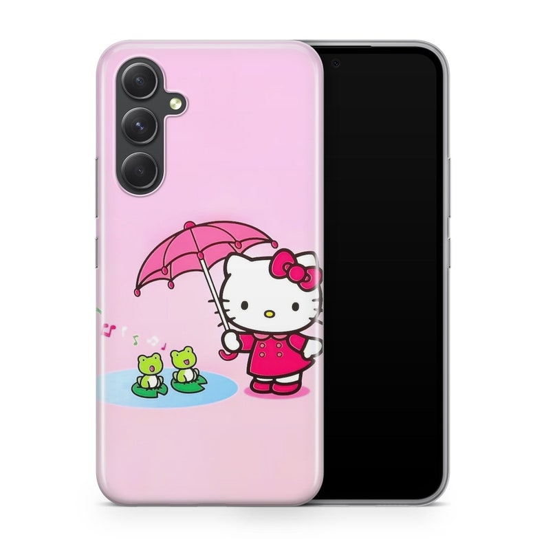 Puede incluir: Una funda de tel&eacute;fono rosa con un dise&ntilde;o de Hello Kitty. Hello Kitty sostiene un paraguas rosa y hay dos ranas verdes en el fondo. La funda del tel&eacute;fono es compatible con un tel&eacute;fono que tiene una lente de c&aacute;mara triple.