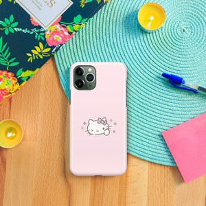 Puede incluir: Una funda de tel&eacute;fono rosa con un personaje Hello Kitty blanco con un lazo rosa sobre un fondo rosa. La funda tiene un recorte para la c&aacute;mara y un contorno blanco alrededor del personaje.
