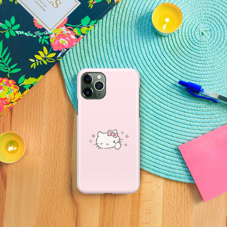 Puede incluir: Una funda de tel&eacute;fono rosa con un personaje Hello Kitty blanco con un lazo rosa sobre un fondo rosa. La funda tiene un recorte para la c&aacute;mara y un contorno blanco alrededor del personaje.