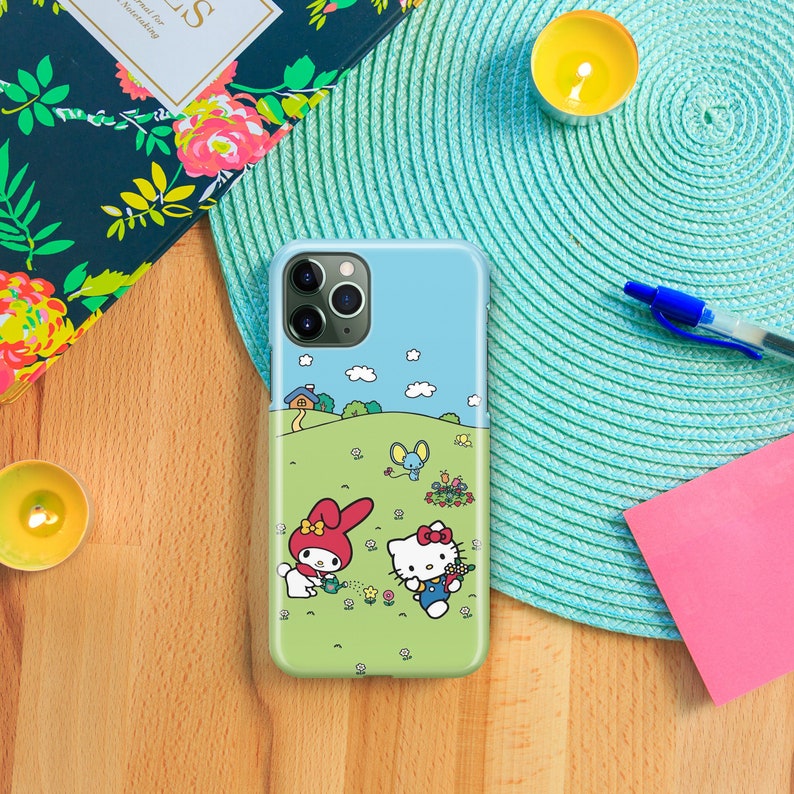 Pu&ograve; includere: Una custodia per telefono blu e verde con un'immagine di cartone animato di Hello Kitty e My Melody che fanno giardinaggio. La custodia ha un ritaglio per l'obiettivo della fotocamera.