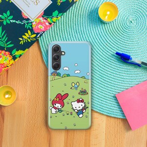 May include: A clear phone case with a blue background and a cartoon image of Hello Kitty and My Melody. The case has a camera cutout and a white border.