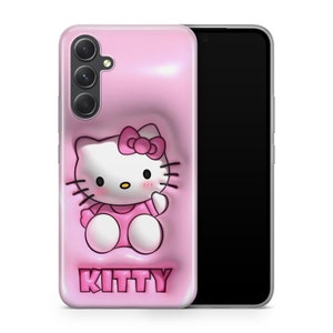 Puede incluir: Una funda de tel&eacute;fono rosa con un dise&ntilde;o de Hello Kitty en 3D. La funda tiene un recorte para el objetivo de la c&aacute;mara y el texto "KITTY" impreso en la parte inferior.