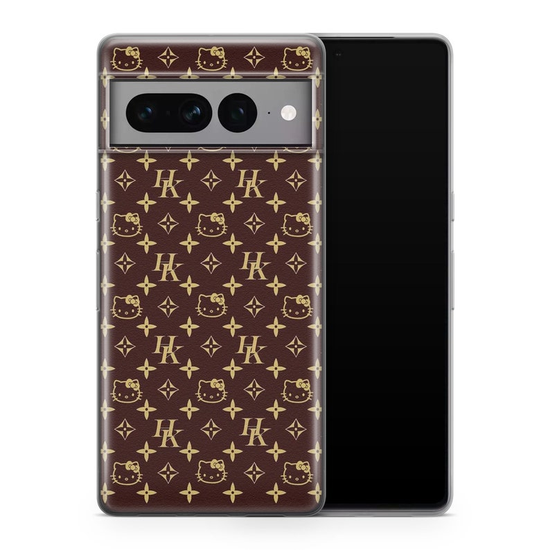 Op de afbeelding: Een transparante telefoonhoes met een bruin en gouden patroon. Het patroon heeft een herhalend ontwerp van een klein wit kattengezicht met een rode strik, een gouden "HK"-monogram en een gouden ruitvorm. De hoes is ontworpen om te passen op een smartphone.