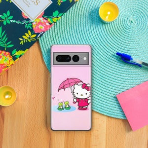 May include: A pink phone case with a Hello Kitty design. The illustration shows Hello Kitty holding a pink umbrella with two green frogs in a pond. The case is on a wooden surface with a teal placemat and a notebook.