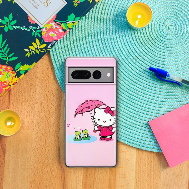 May include: A pink phone case with a Hello Kitty design. The illustration shows Hello Kitty holding a pink umbrella with two green frogs in a pond. The case is on a wooden surface with a teal placemat and a notebook.