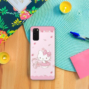Op de afbeelding: Een roze telefoonhoesje met een Hello Kitty-ontwerp. Hello Kitty houdt een roze teddybeer vast. Het hoesje heeft een roze achtergrond met witte bloemen en aardbeien.